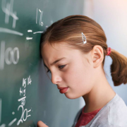 Seorang siswa perempuan berdiri di depan papan tulis dengan dahi menyentuh permukaannya, tampak kesulitan memahami soal matematika berupa pecahan dan pembagian.