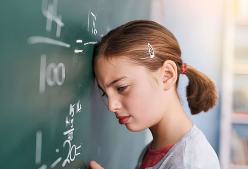 Seorang siswa perempuan berdiri di depan papan tulis dengan dahi menyentuh permukaannya, tampak kesulitan memahami soal matematika berupa pecahan dan pembagian.