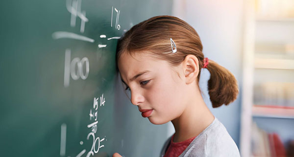 Seorang siswa perempuan berdiri di depan papan tulis dengan dahi menyentuh permukaannya, tampak kesulitan memahami soal matematika berupa pecahan dan pembagian.